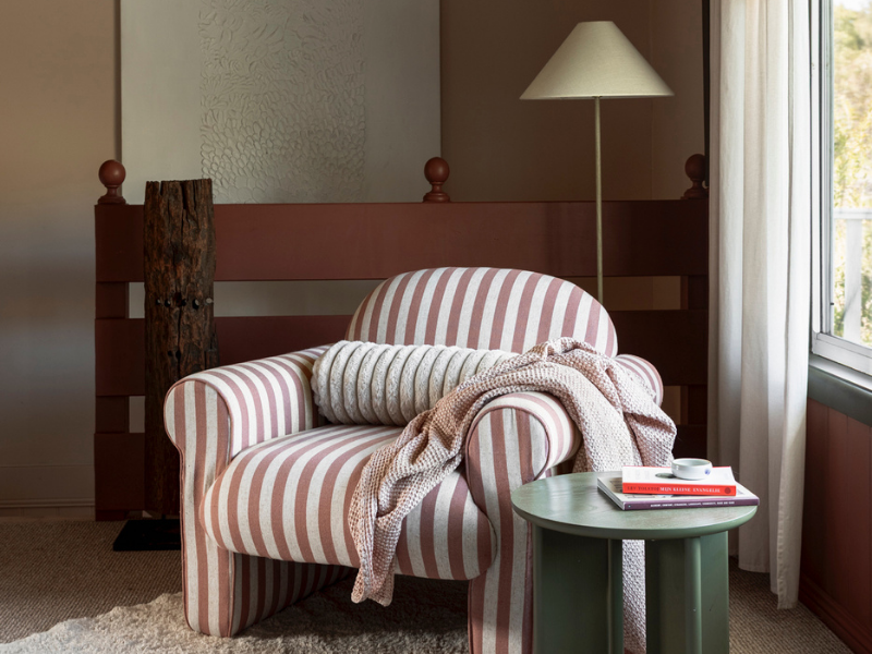 Cozy armchair with red and white stripes, draped with a knitted blanket and round pillow. Nearby, a small green table with books and a cup, under soft lighting.