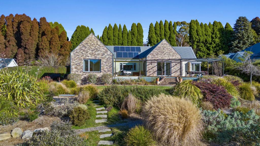A stone house with solar panels on the roof is surrounded by lush greenery and a bright garden. Tall trees stand in the background under a clear blue sky. Located at 40 Hoddle Street, Burrawang.