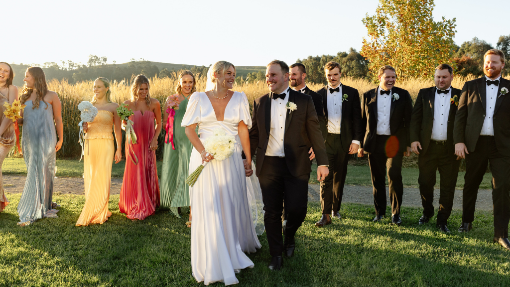 A joyful wedding party walks on grass with a bride in a white gown and groom in a black suit center. Bridesmaids wear colorful dresses, groomsmen in suits.