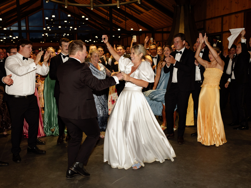A joyous bride in a white gown dances with a groom in a black suit, surrounded by cheering guests in formal attire. The atmosphere is festive and lively.
