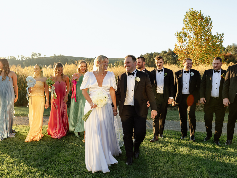 A joyful wedding party walks on grass under a sunny sky. The bride in a flowing white gown holds a bouquet, and the groom is in a black tuxedo. Bridesmaids in colorful dresses, and groomsmen in tuxedos, surround them, smiling amid tall fields and autumn trees.