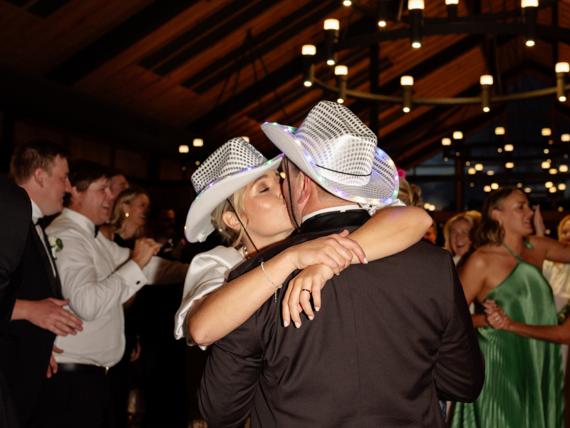 A couple wearing glowing cowboy hats shares a kiss on a dance floor. They're surrounded by smiling guests in formal attire, under warm overhead lights.