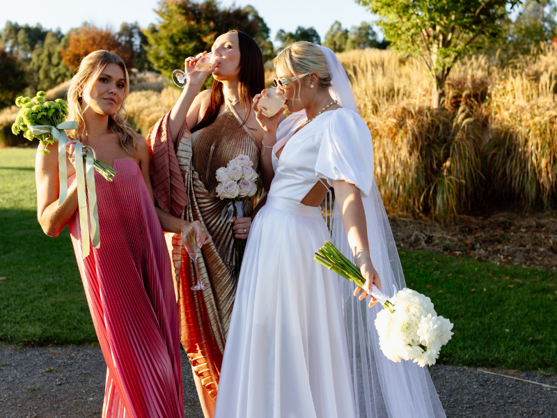Three women in elegant dresses enjoy drinks outdoors. The bride in white, holding flowers, stands beside two bridesmaids in colorful gowns. Joyful and relaxed.