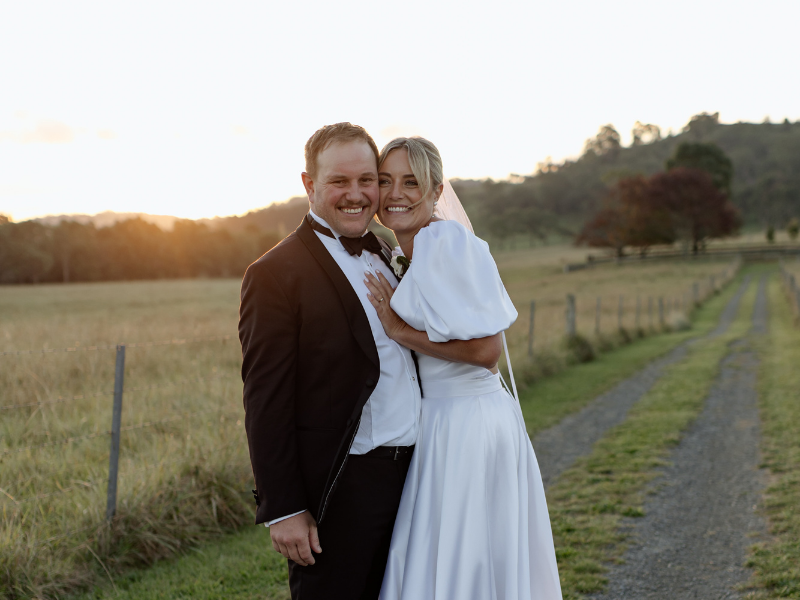 A bride in a white dress and groom in a black suit smile joyfully at sunset on a country path, with rolling fields and trees in the background.