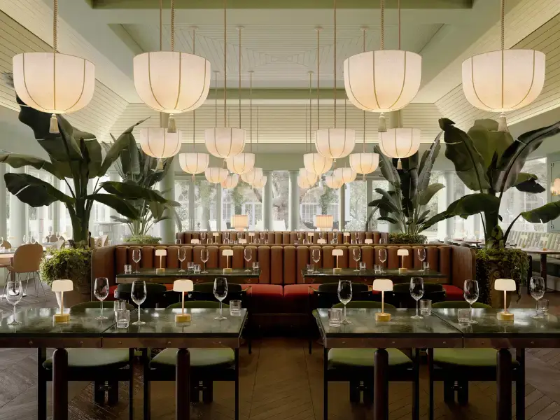 Elegant restaurant interior with green walls, ambient lighting from large hanging lamps. Tables set with wine glasses and small lamps, surrounded by lush plants.