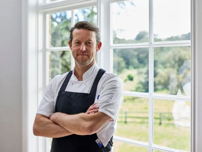 Chef in a crisp white shirt and black apron stands confidently with arms crossed, in front of a bright window showing a lush, green landscape.