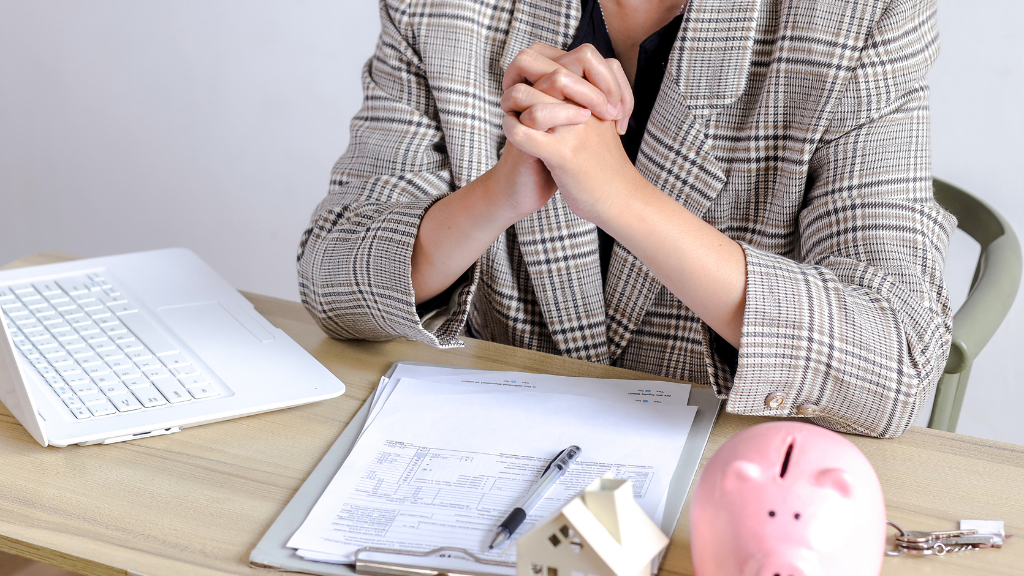 Person in plaid blazer sits at a desk with hands clasped. Papers, a pen, a small house model, and a pink piggy bank are on the table, suggesting planning.