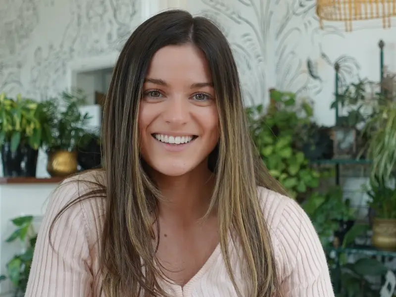 Smiling woman with long brown hair wearing a light pink sweater, seated indoors. The background features green plants and patterned wallpaper, creating a cozy, natural ambiance.