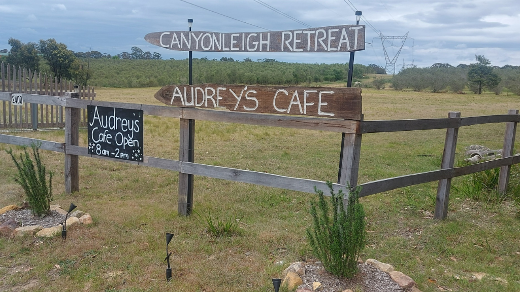 A rustic wooden sign for "Canyonleigh Retreat" and "Audrey's Cafe" stands on a grassy field under a cloudy sky. The cafe is open from 8 AM to 2 PM.
