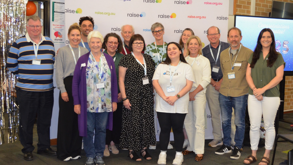 A group of 14 people stands smiling in front of a "raise.org.au" backdrop. They are diverse in age and attire, conveying a joyful and celebratory atmosphere.