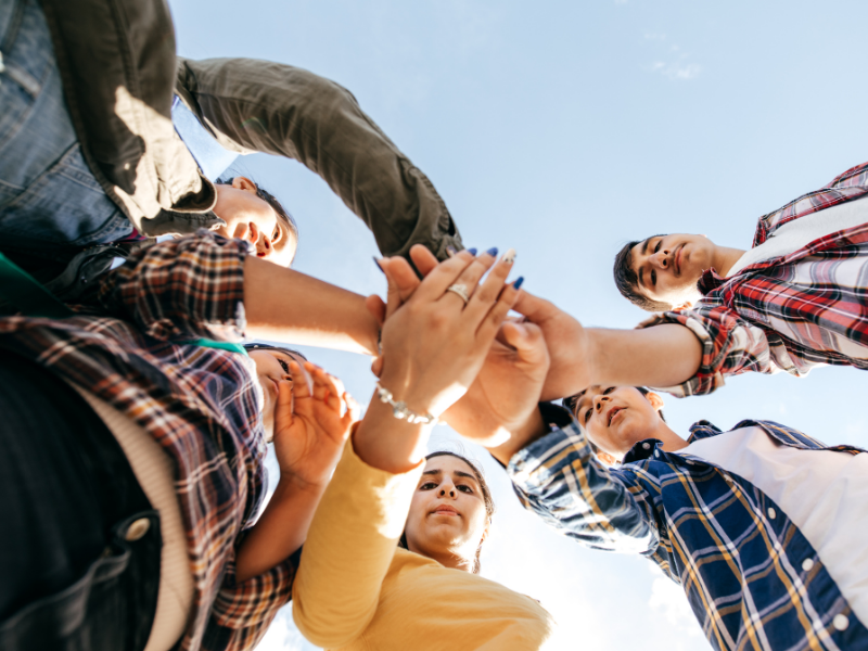 A diverse group of people stand in a circle, reaching their hands towards the center in a gesture of unity. Bright sky in the background.