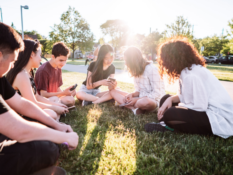 A group of young people sits in a circle on grass in a sunlit park, engaging with their smartphones and each other, conveying a cheerful and relaxed atmosphere.