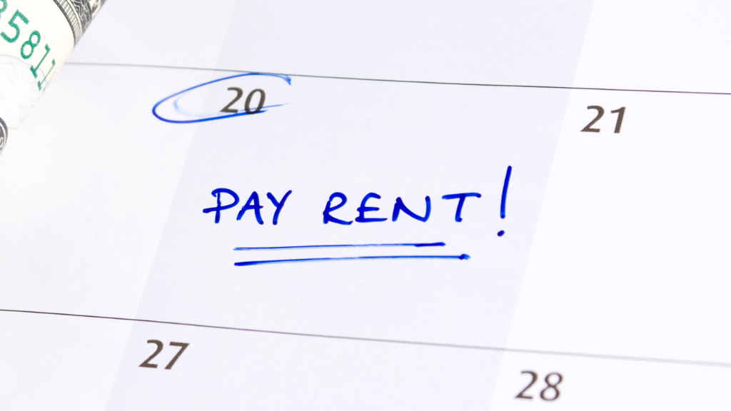 Calendar page with the 20th circled in blue and "PAY RENT!" written prominently. A dollar bill is partially visible on the left. Reminder and urgency conveyed.
