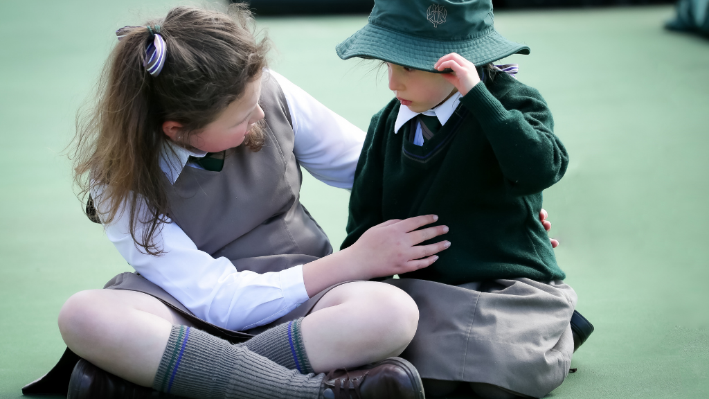 Two children, seated on the ground, share a comforting moment. One gently supports the other, wearing a green sweater and hat, conveying warmth and empathy.