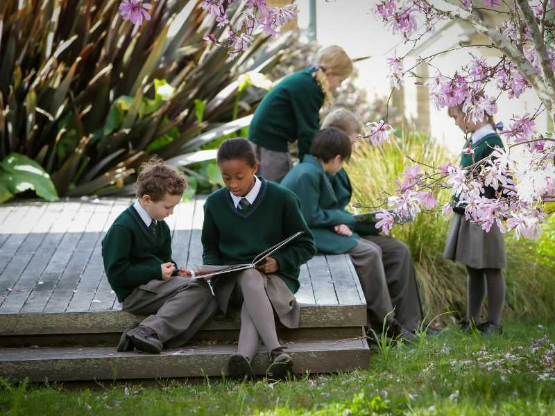 Children in green uniforms read outdoors on wooden steps surrounded by lush plants and pink blossoms. The scene is calm and focused.