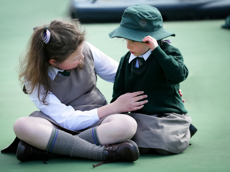 A young girl in a school uniform, sitting on a green floor, comforts a younger child wearing a green sweater and bucket hat. She appears supportive and caring.