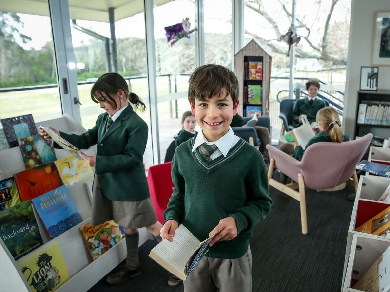 Smiling boy in school uniform holds an open book in a bright library. Other children read in the background. Atmosphere is warm and studious.