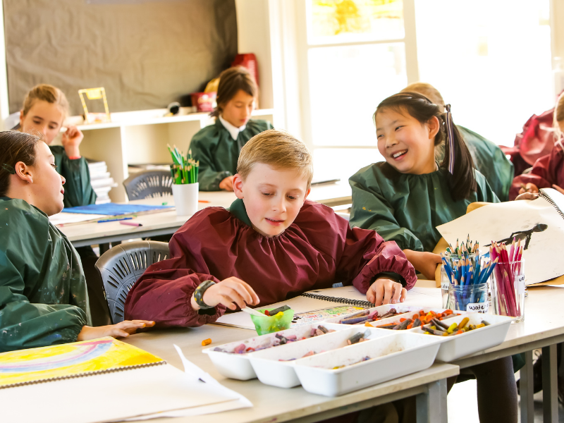 Children wearing smocks sit around a table, engaged in drawing with colorful pencils. The room is bright, fostering a lively, creative atmosphere.