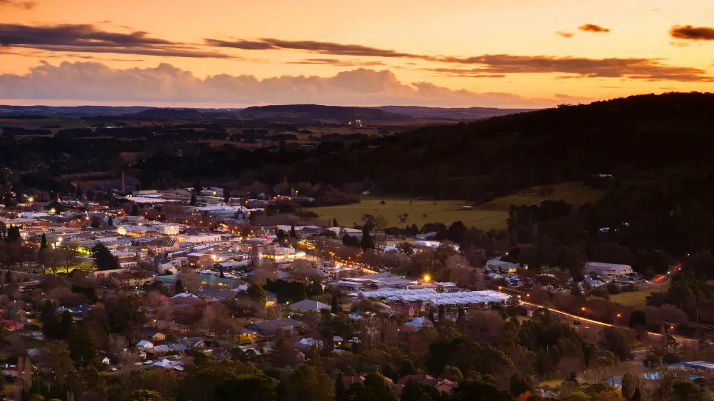 Aerial view of a town at sunset, with warm lights illuminating buildings and streets. Dark hills border the scene under a soft, colorful sky.