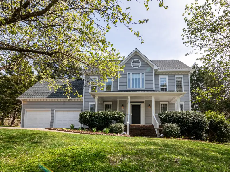 A charming two-story gray house with white trim, featuring a welcoming front porch and well-kept shrubs, sits under a leafy tree on a sunny day.
