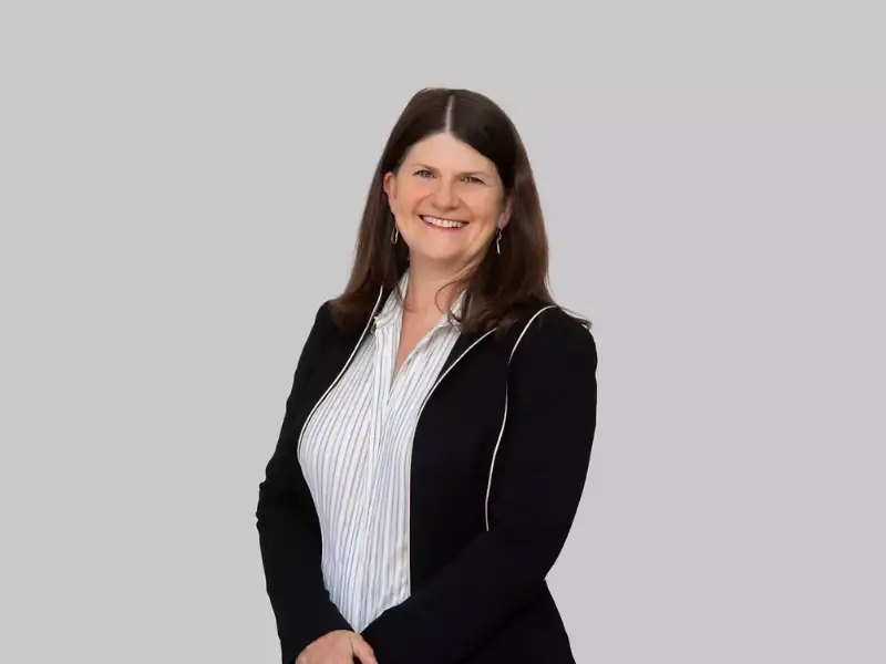 Smiling woman with long brown hair stands against a light gray background, wearing a black jacket over a striped shirt, conveying professionalism.