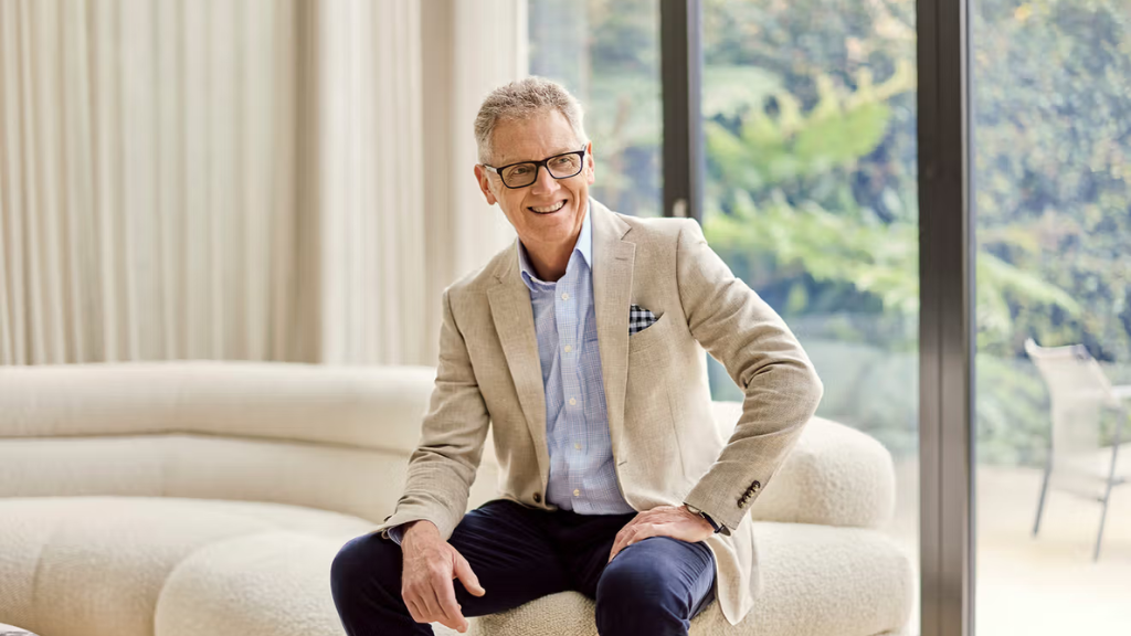 A man in glasses, wearing a beige blazer and blue shirt, sits on a cream sofa in a bright, modern room with large windows and lush greenery outside.