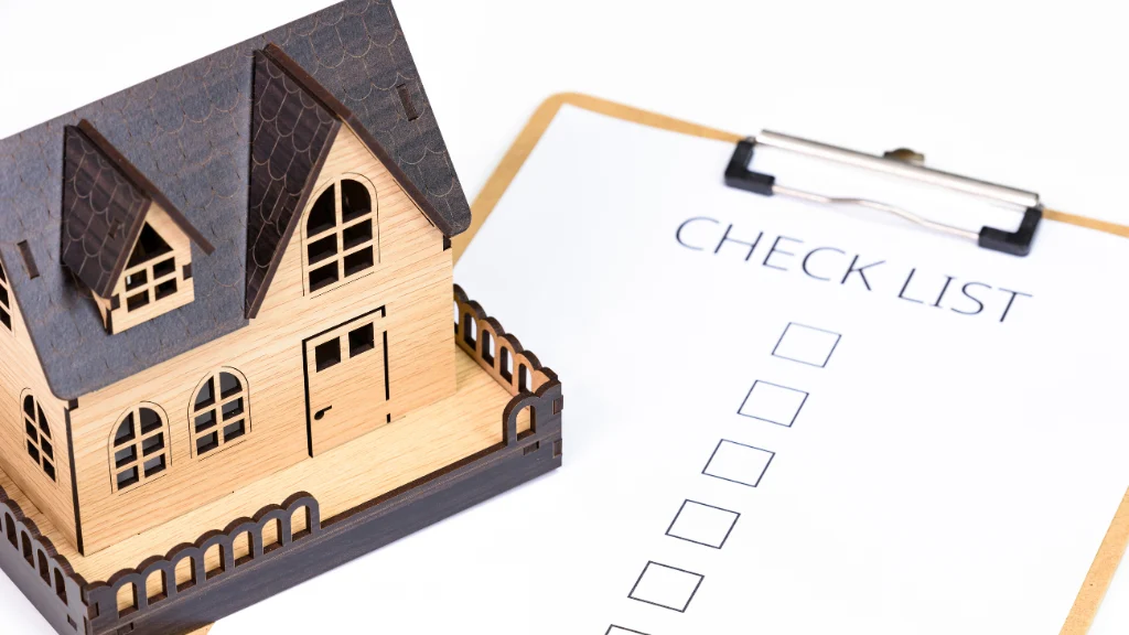 A wooden house model sits on a table beside a checklist on a clipboard. The checklist is blank, suggesting tasks or items for house planning.