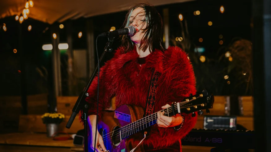 female singer in red jacket singing into a microphone and holding a guitar